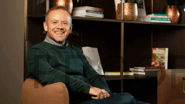 a man in a green sweater sits in front of a bookshelf with books on it