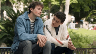 two young men are sitting on a bench in a park talking .