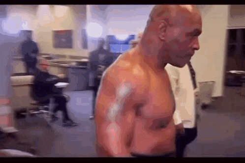 a man without a shirt is standing in a room with a man sitting in a chair in the background .