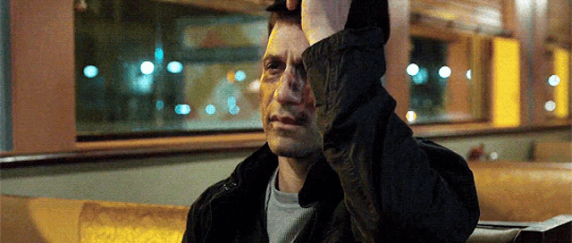 a man in a black jacket is sitting on a couch covering his face with his hand .