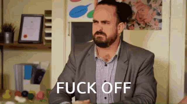 a man in a suit and plaid shirt is sitting in front of a sign that says " fuck off "