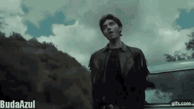 a man in a leather jacket is standing next to a car in a dark forest .