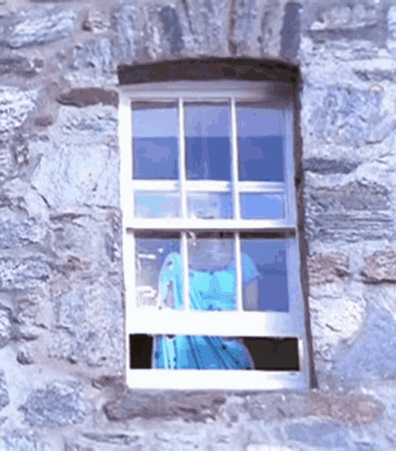 a woman in a blue dress is looking out of a window in a stone wall