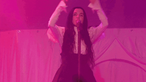 a woman is singing into a microphone with her arms in the air in front of a pink curtain .