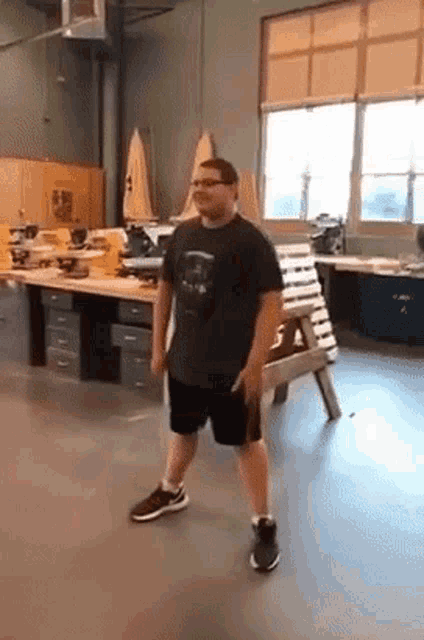 a man in a black shirt and shorts is standing in a room with a wooden bench in the background .