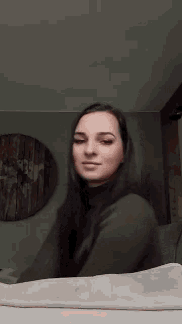 a woman in a green sweater is sitting on a bed in front of a wall clock .