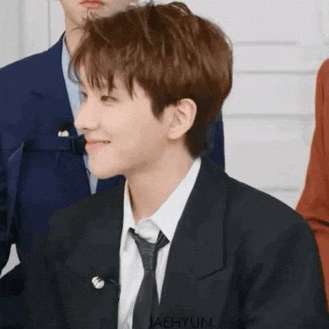 a young man in a suit and tie is smiling while sitting in front of a group of people .