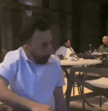 a man in a blue shirt is sitting at a table in a restaurant talking on a cell phone .