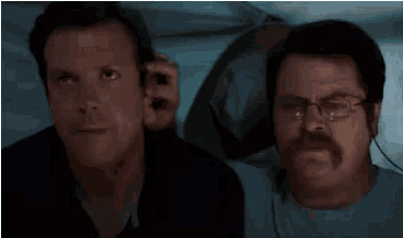 two men are sitting next to each other in bed . one of the men is wearing glasses and a mustache .
