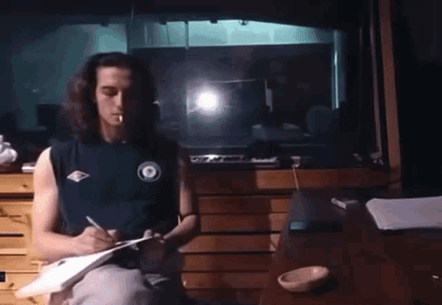 a man with long hair is sitting at a desk writing on a piece of paper and smoking a cigarette