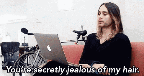 a man with long hair is sitting on a couch using an apple laptop and saying you 're secretly jealous of my hair
