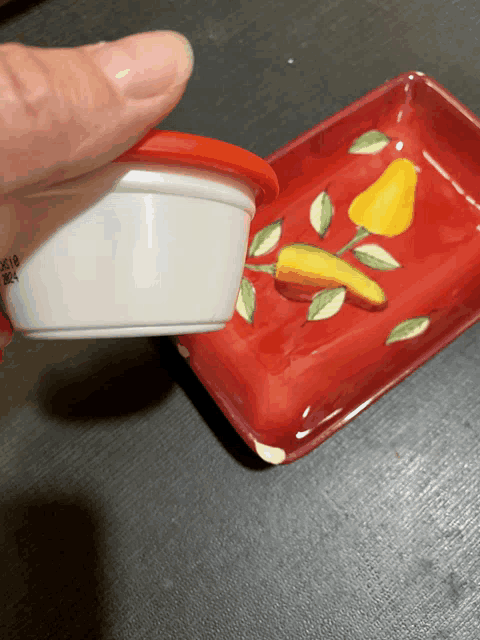 a person is holding a small white container with a red lid over a red plate with lemons on it