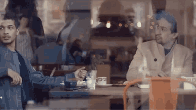 two men are sitting at a table in a restaurant and one of them is pointing