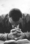 a man in a suit and tie is sitting at a table with his hands folded in prayer over a bible .