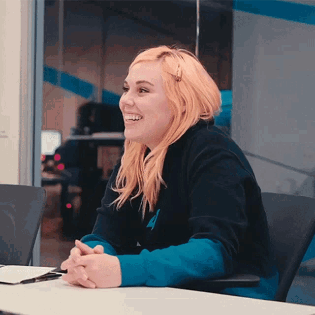 a woman with pink hair is smiling while sitting at a desk with her hands folded