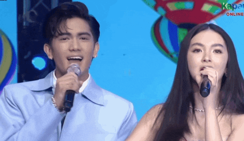 a man and a woman singing into microphones in front of a blue background that says kapa online