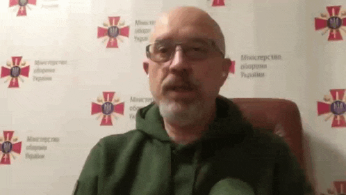 a man with a beard and glasses is sitting in a chair in front of a wall with a cross on it .