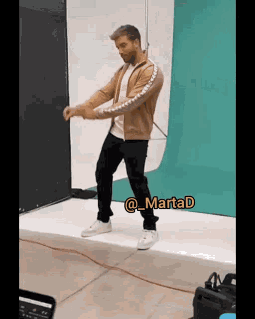 a man in a tan jacket and black pants is dancing in front of a green and white backdrop with the hashtag martad