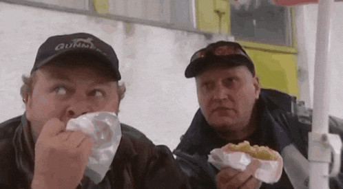 two men are sitting at a table eating hot dogs . one of the men is wearing a gunn 's hat .