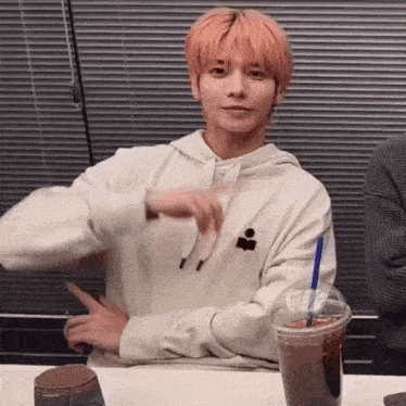 a young man with pink hair is sitting at a table with a cup of coffee and making a funny face .