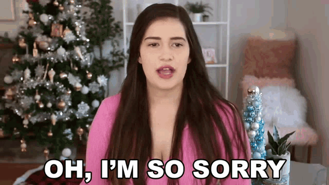 a woman in a pink sweater is standing in front of a christmas tree and says oh , i 'm so sorry