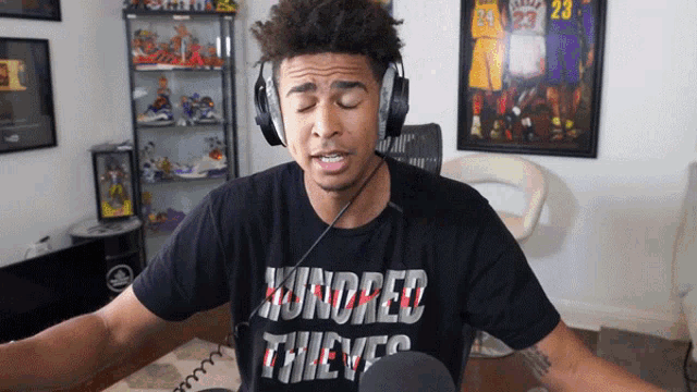 a man wearing headphones and a hundred thieves shirt stands in front of a microphone