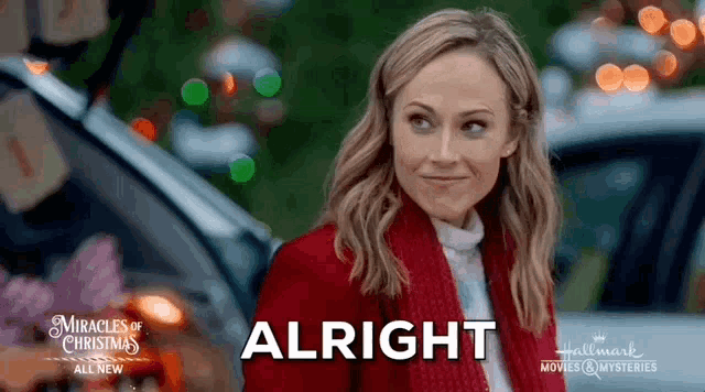 a woman in a red coat is standing in front of a car with the word alright on her face .