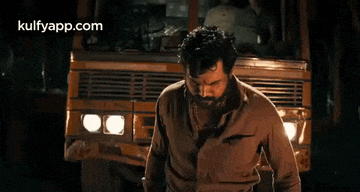 a man with a beard is standing in front of a yellow truck and looking at the camera .