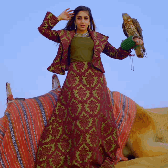 a woman in a long dress holds a bird in her hand while standing next to a camel
