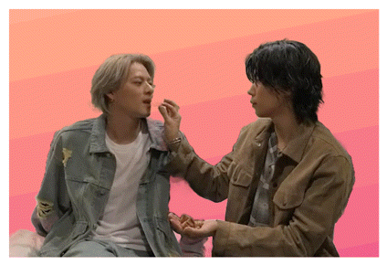 two men are sitting next to each other on a pink background and one of them is feeding the other .