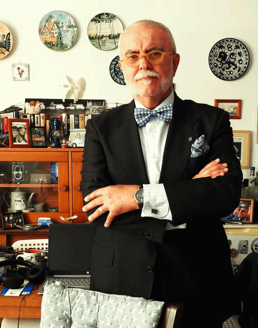 a man in a suit and bow tie stands with his arms crossed in front of a laptop