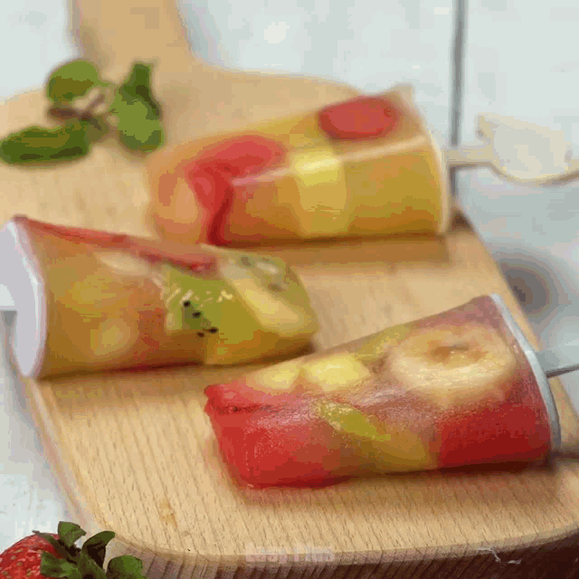three fruit popsicles are on a wooden cutting board with a strawberry in the background