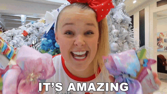 a girl with a red bow on her head says it 's amazing in front of a christmas tree
