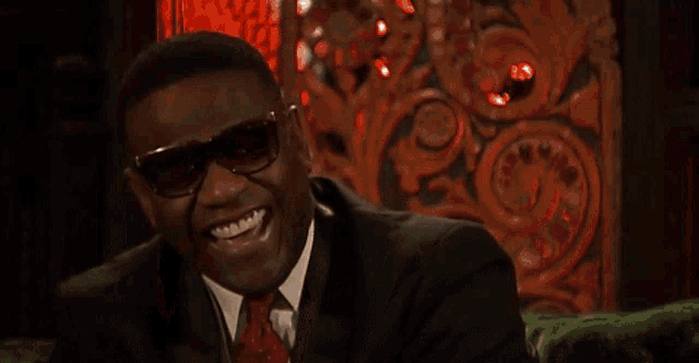 a man wearing sunglasses and a suit is smiling in front of a red curtain