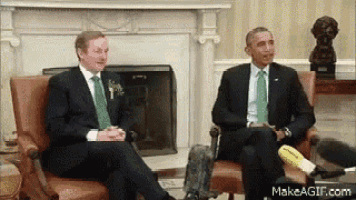 two men in suits and green ties are sitting in chairs in front of a fireplace on makeagif.com