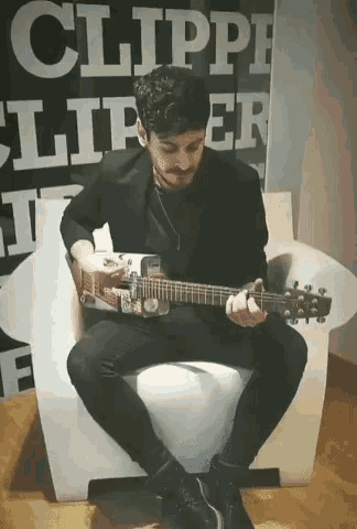 a man is sitting in a chair playing a guitar in front of a sign that says clipper