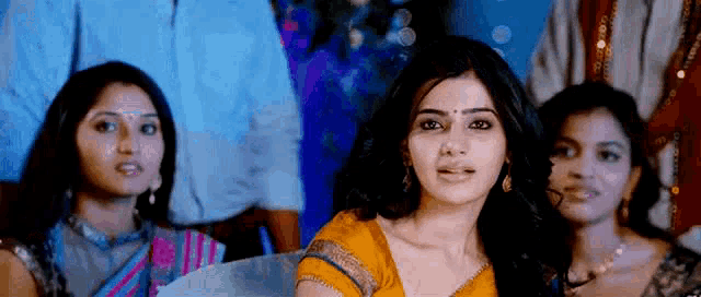 a group of women are sitting in a room and one of them is wearing a yellow saree .