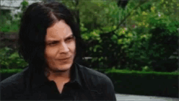 a man with long black hair and a black shirt is standing in front of a hedge .