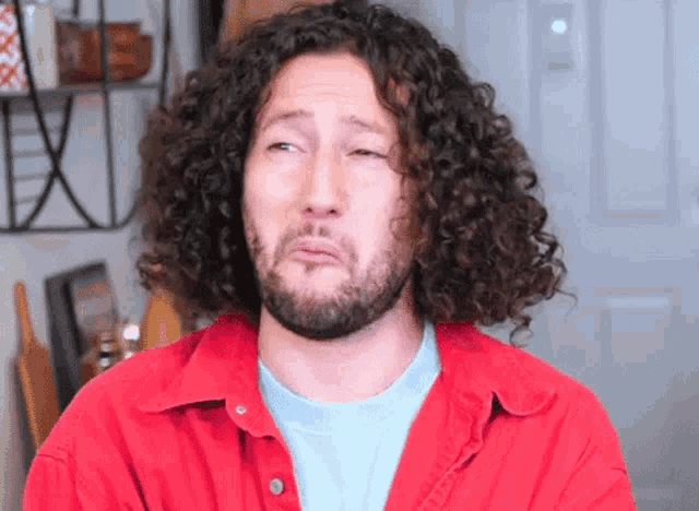 a man with curly hair and a beard is wearing a red shirt .