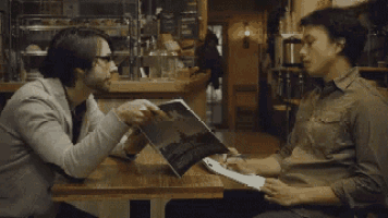 two men are sitting at a table in a restaurant and one is holding a clipboard