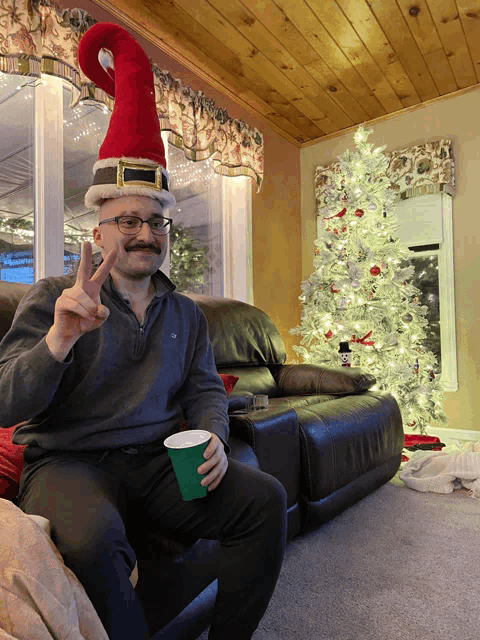 a man wearing a santa hat is sitting in front of a christmas tree and giving the peace sign
