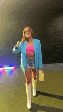 a woman wearing a blue blazer , pink crop top , denim shorts and white boots is standing on a street .