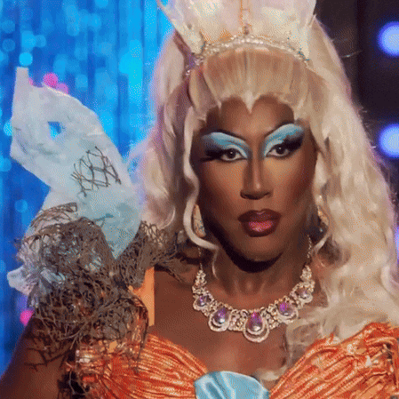 a drag queen wearing a crown and a necklace looks at the camera