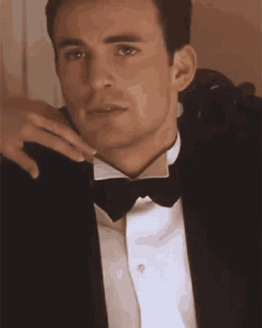 a man in a tuxedo and bow tie is sitting in a chair with his hand on his shoulder .