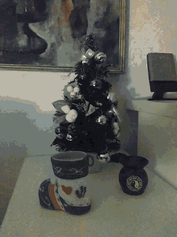 a small christmas tree sits on a table next to a cup and a painting