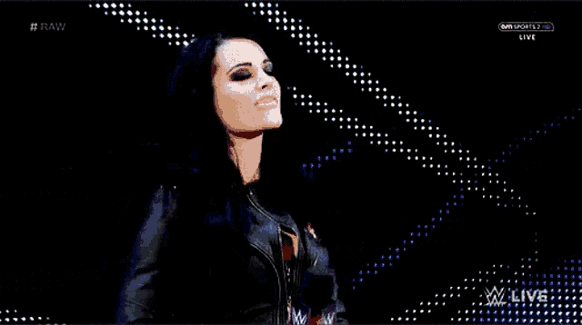 a woman in a black leather jacket is standing in front of a screen that says raw
