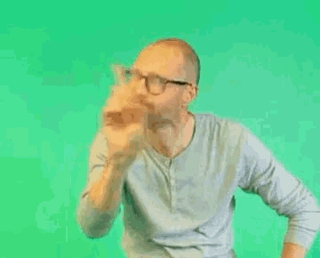 a man wearing glasses and a gray shirt is standing in front of a green screen .