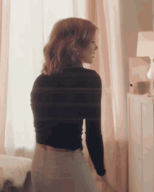 a woman in a black top stands in front of a window with pink curtains