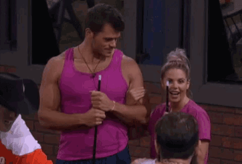 a man in a pink tank top is standing next to a woman in a pink shirt and holding a wand .