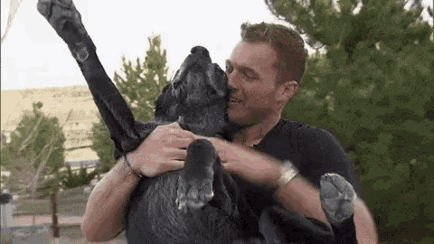 a man is holding a black dog in his arms and the dog is licking the man 's face .